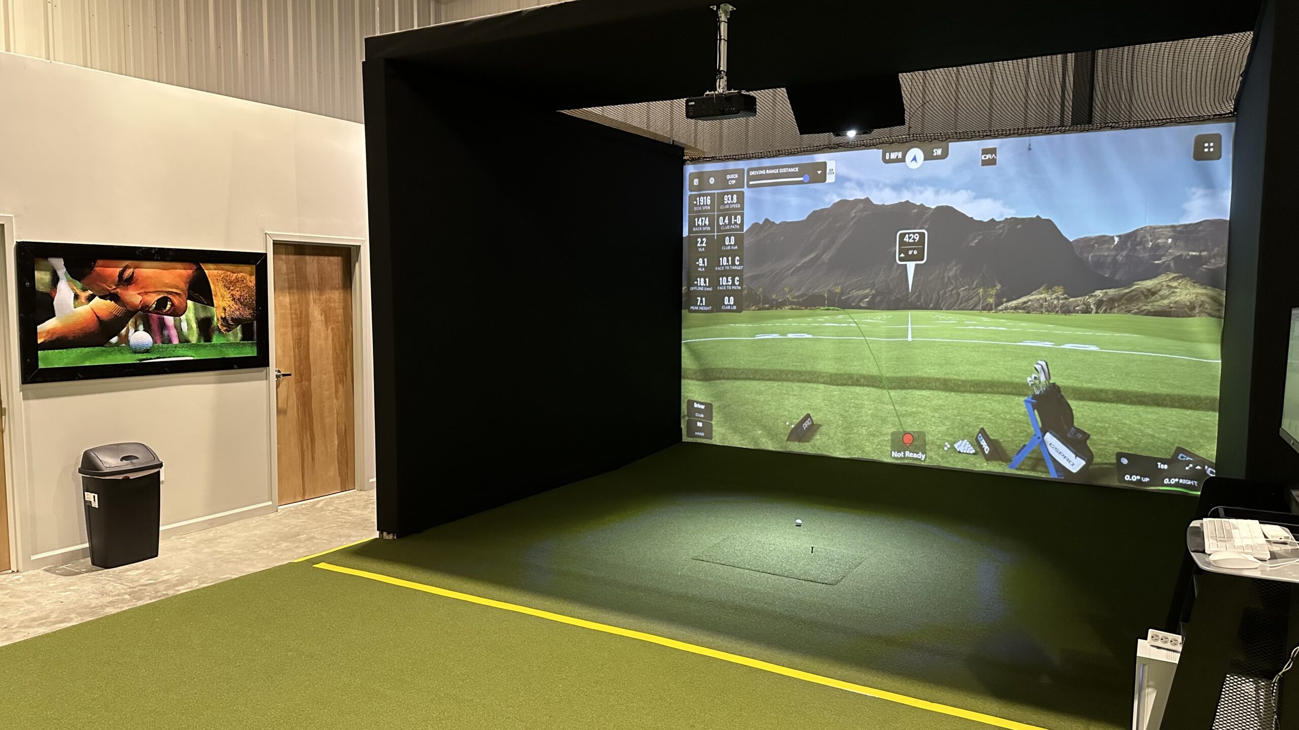 Indoor golf simulator setup with a large screen displaying a golf course, artificial turf flooring, a golf ball, and a TV on the wall—all enhanced by seamless Tee Time Booking through Tee24 for the ultimate golfing experience.