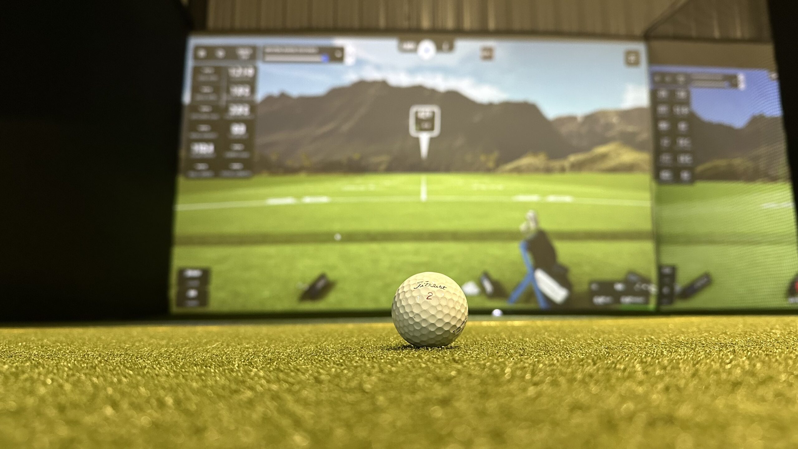 A golf ball sits on artificial turf in front of a large virtual golf simulator screen displaying a golf course—perfect for practicing before your next round with easy tee time booking through Tee24.