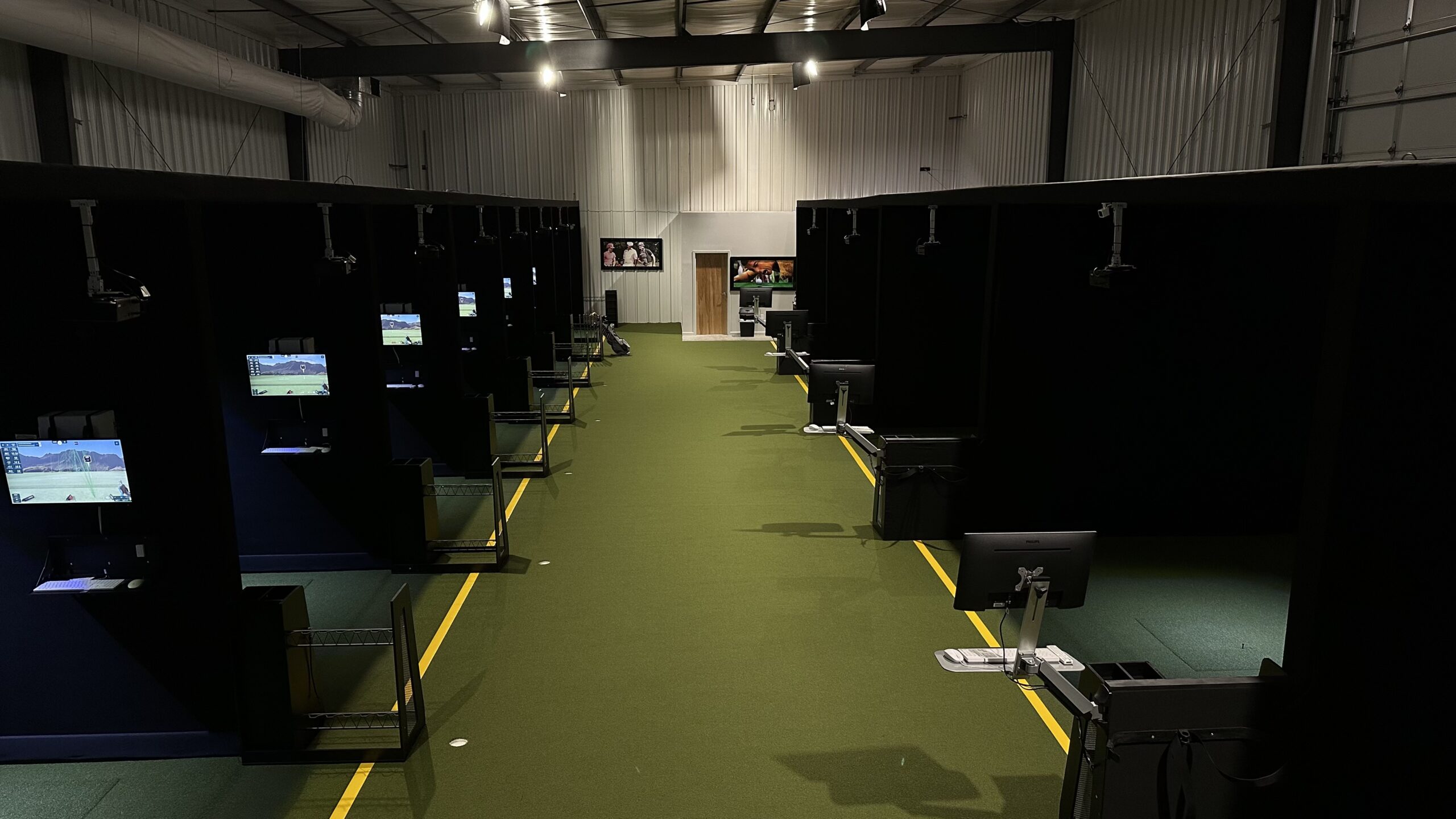 Tee24 offers an indoor golf simulator facility with several individual booths, each equipped with a screen and equipment, all lined up along a green artificial turf floor—perfect for easy tee time booking.
