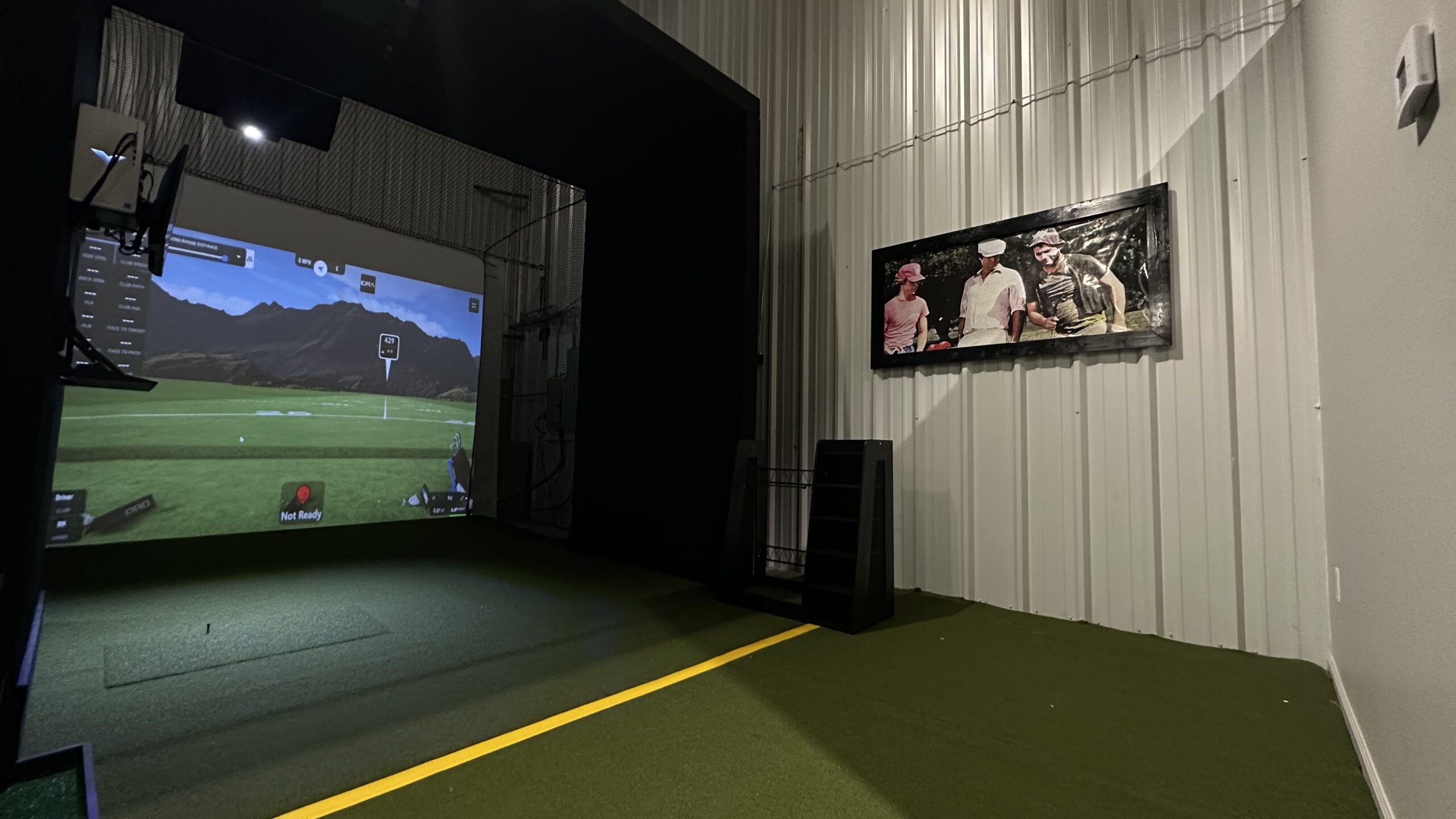 Indoor golf simulator setup featuring a realistic projected golfing scene and a framed photo of golfers on the corrugated metal wall—perfect for your next Tee24 Golf Reservation or tee time booking.