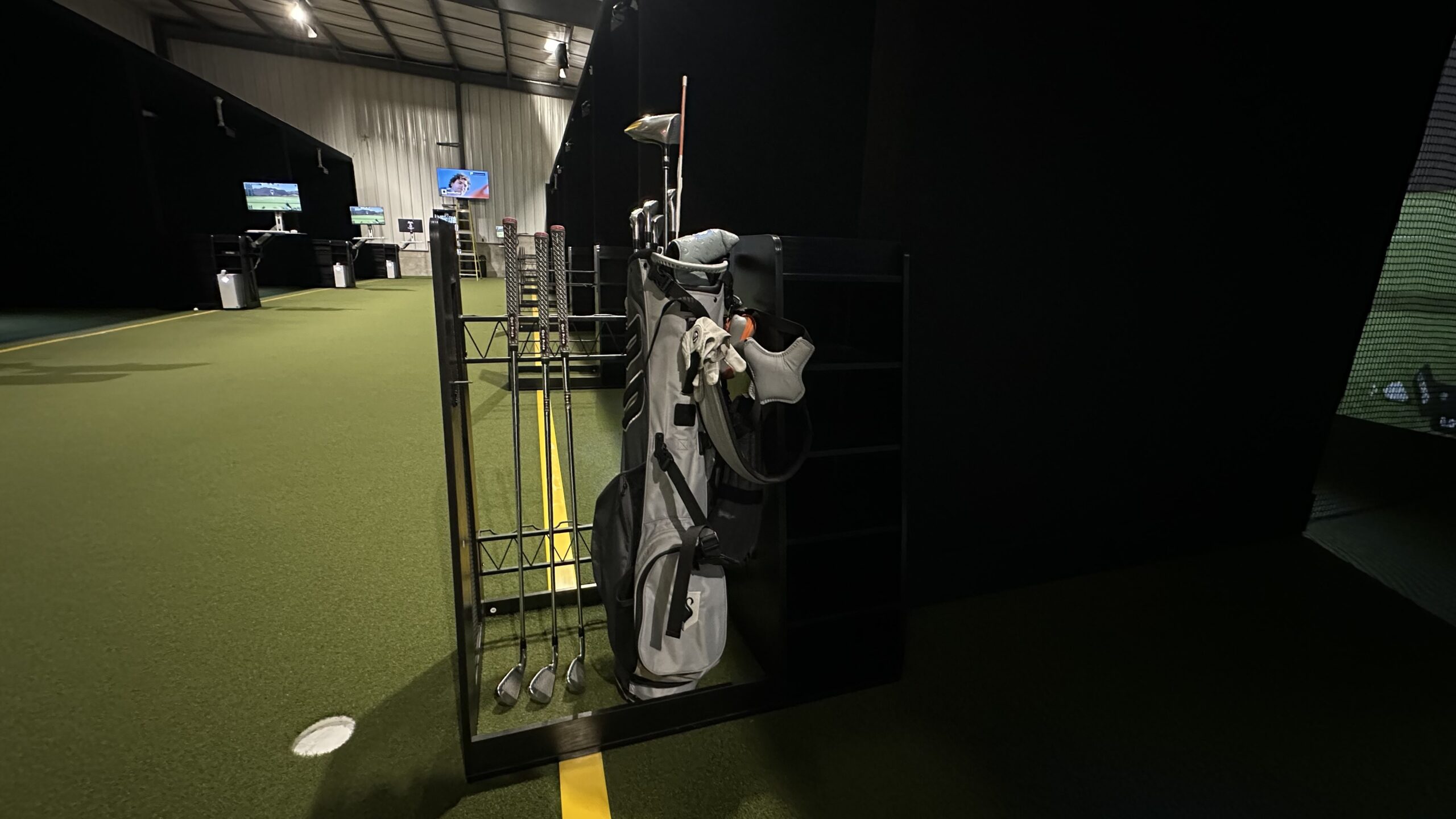 A golf bag and several golf clubs are stored in a rack inside an indoor golf facility with artificial turf and practice areas, offering convenient Tee Time Booking through Tee24.