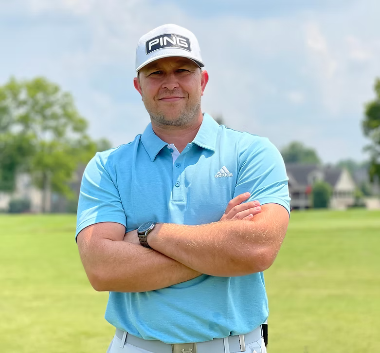 Man wearing a light blue Adidas polo, PING cap, and watch stands with arms crossed on a golf course with trees and houses in the background, ready for Tee24 lessons.